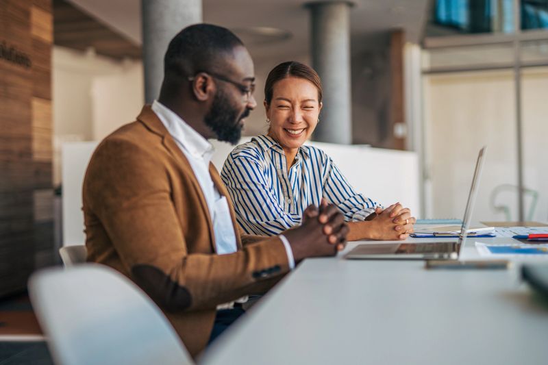 Diverse business colleagues collaborating and smiling while working together on a laptop at a table in a modern office, enjoying a productive and positive work environment