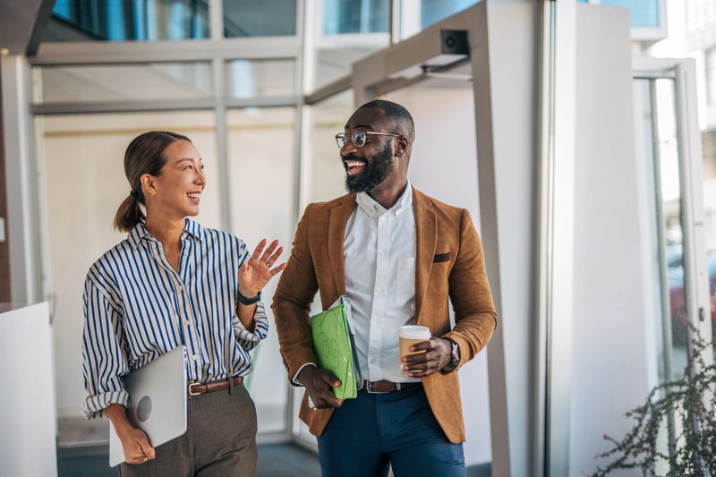 Two diverse business colleagues walk and chat through a modern office, smiling and carrying a laptop, documents, and coffee as they discuss work and collaborate confidently
