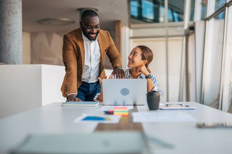Multiracial adult business colleagues are collaborating and smiling while working together on a laptop at a desk in a spacious, contemporary office environment