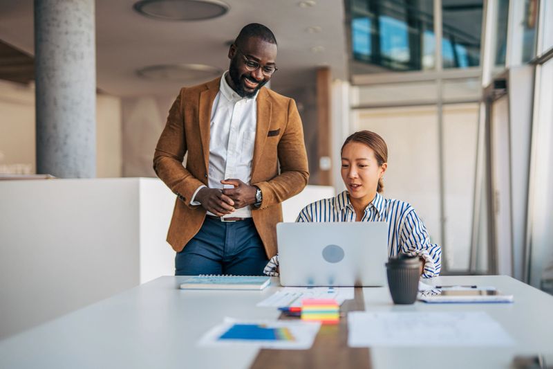 Diverse business colleagues reviewing data on a laptop, discussing work at a desk in a contemporary office setting, highlighting teamwork and productivity