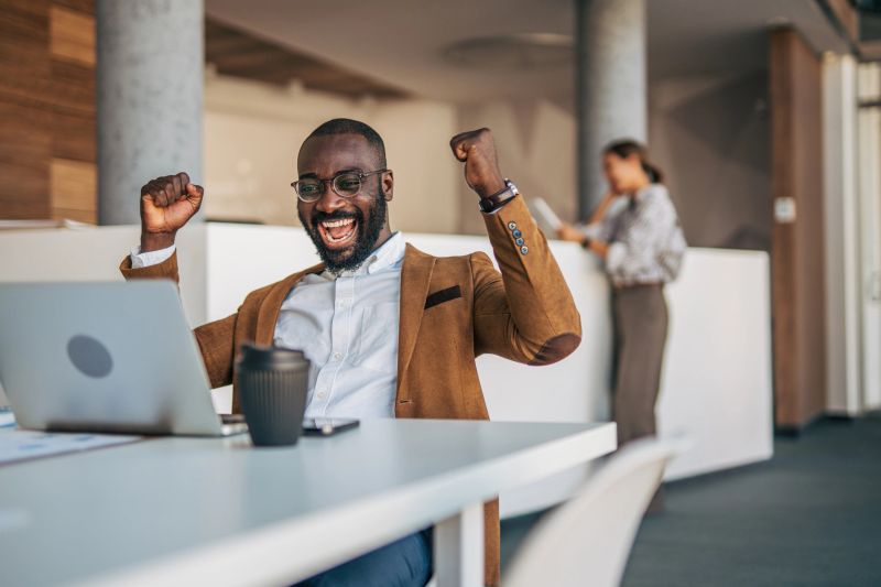 Black businessman celebrating at his laptop with raised fists in a modern open-plan office while a colleague works in the background, showing joy and professional success