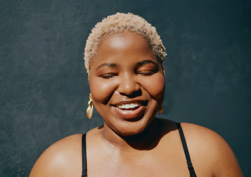 Close-up of a joyful young Black woman with short blonde hair smiling with closed eyes against a dark background, wearing gold earrings and a black top in bright natural light.