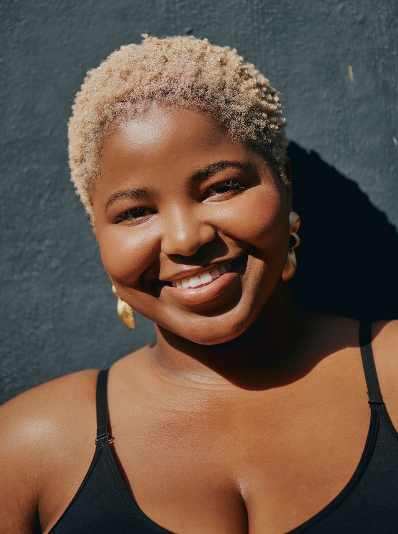 Portrait of a joyful young Black woman with short curly blonde hair wearing gold earrings and a black tank top, smiling warmly against a dark textured background in natural light.