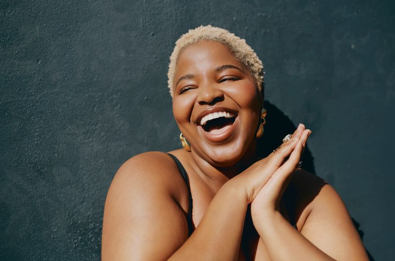 Cheerful young Black woman with short blonde hair smiling broadly and clasping hands in front of a dark textured wall, radiating happiness and confidence in natural daylight.