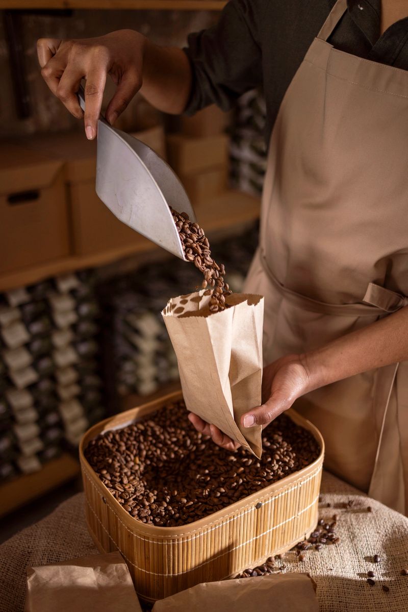 A barista in an apron pours roasted coffee beans into a brown paper bag from a metal scoop, over a wooden bin in a warm, rustic cafÃ© or roastery scene