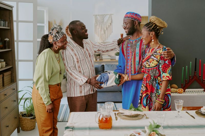 Group of Black adults standing around table celebrating Kwanzaa, smiling and exchanging wrapped gift, kinara with candles visible on table, traditional clothing worn by participants