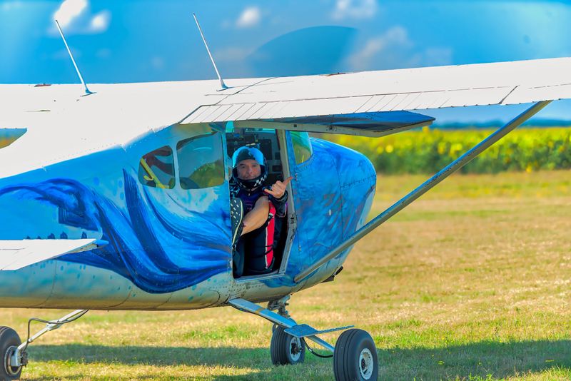 A pair of skydivers sit in a compact airplane, buckled and ready as the open door awaits exit. One jumper playfully signals a phone call to ground crew, capturing a calm, focused moment of teamwork, safety checks, and anticipation before ascent and jump.
