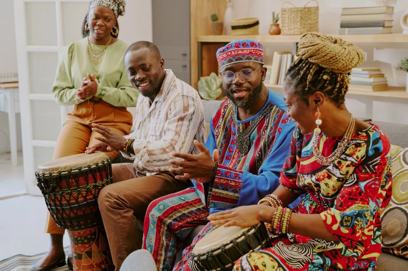 Group of Black adults celebrating Kwanzaa, sitting together on sofa, playing traditional drums, smiling and engaging in conversation, woman standing clapping hands nearby