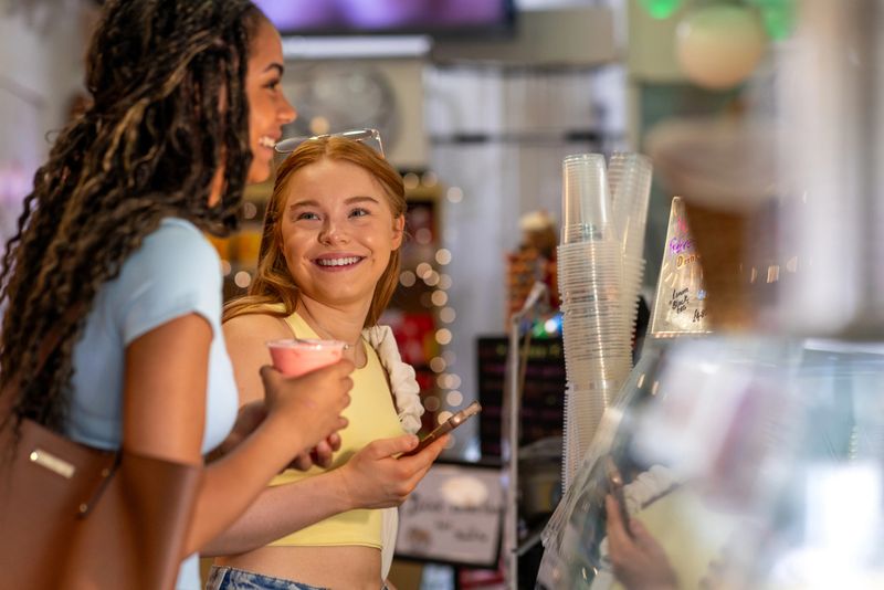 A medium side view of two smiling young women enjoying their time together at a bubble tea shop in the North East of England. They stand at the counter, warmly lit with fairy lights. The cheerful atmosphere, decorated with fairy lights and posters, highlights the welcoming vibe of this local café. Perfect for concepts of friendship, leisure, modern payment, and socialising.Videos are available similar to this scenario.