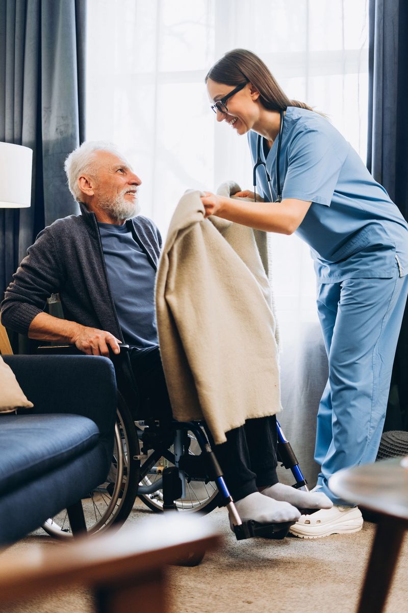 Smiling nurse covering a senior man using a blanket in his wheelchair, showing care and assistance in home environment