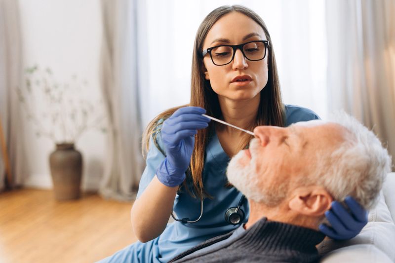 Female healthcare worker in uniform and gloves performing a COVID-19 nasal swab test on a mature adult man during a home visit