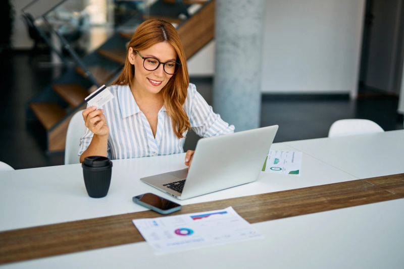 Young woman shopping online at her office desk, smiling as she enters credit card details on a laptop, embracing modern e-commerce and secure digital payments