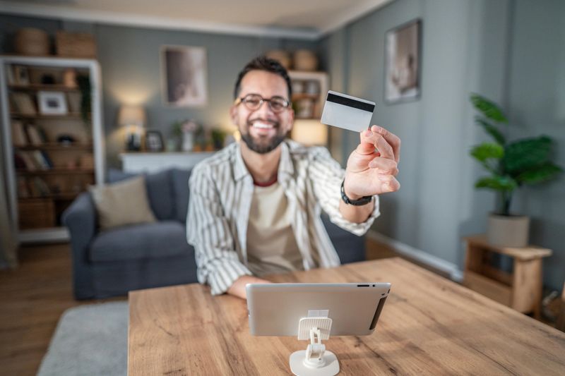 Cheerful man holding a credit card, confidently making an online payment or transaction using a digital tablet at home, symbolizing secure and convenient e-commerce