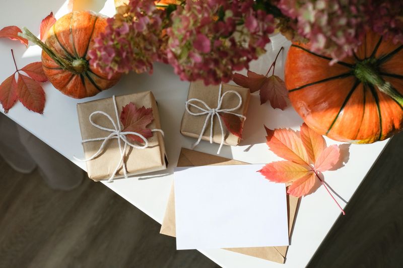 Cozy autumn flat lay with two wrapped gifts, pumpkins, fall leaves and an empty greeting card on white surface. Perfect for seasonal celebrations and Thanksgiving