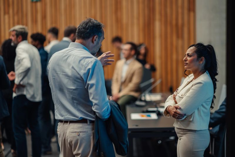 Two business professionals networking and having a serious conversation at a professional event with other blurred colleagues in the background