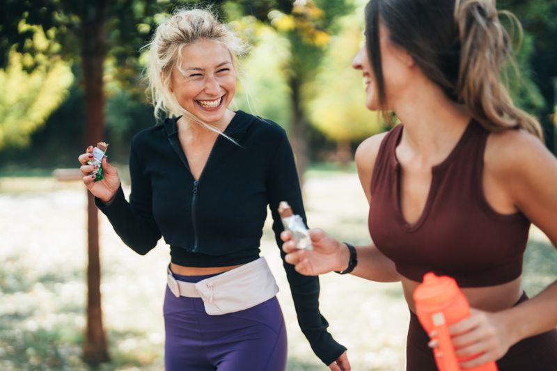 Two fit friends walking together in a park after exercising, smiling and enjoying protein healthy bars. The scene captures post-workout camaraderie and the importance of nutrition and friendship in fitness routines.
