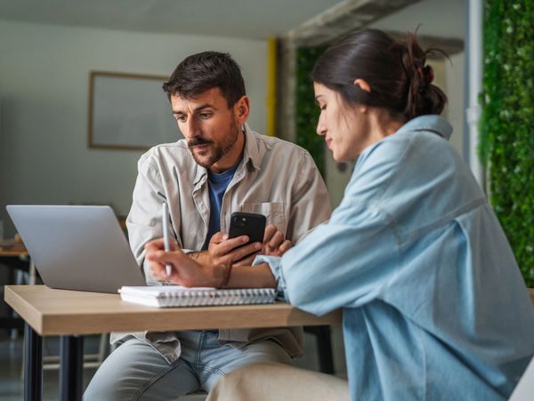 Two colleagues discussing work at a table with a laptop and notebook.