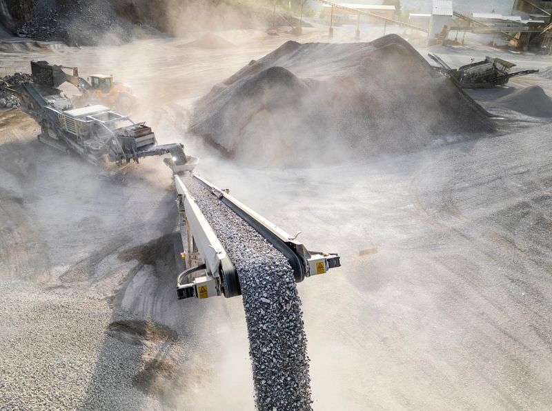 An industrial gravel conveyor moves rocks in a dusty quarry. Heavy machinery operates nearby, surrounded by piles of gravel under soft daylight.