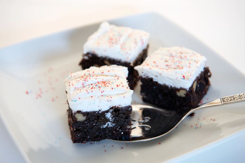 Select focus of squares of sprinkled, frosted, dark chocolate brownies served on white, square plate.