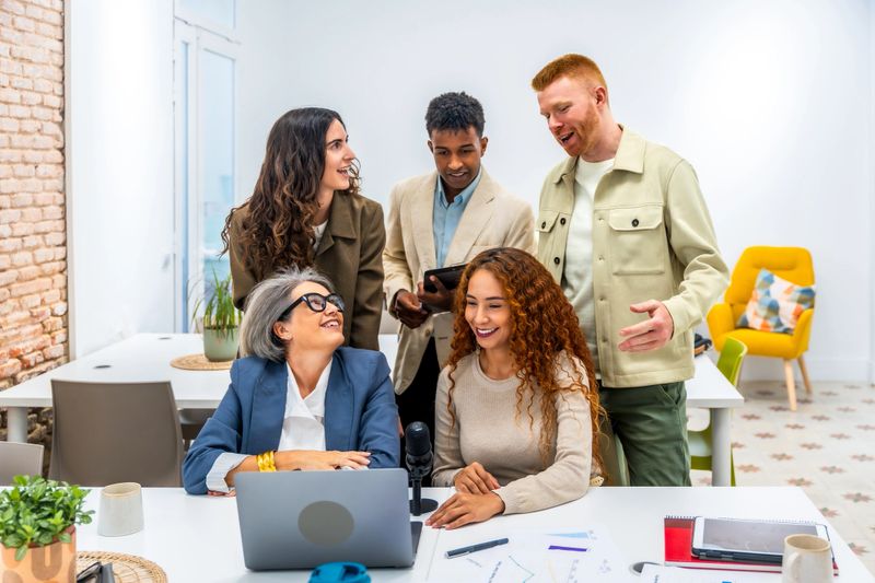 Diverse group of smiling colleagues collaborating on a podcast episode in a bright, modern office, discussing ideas and using a laptop and microphone while recording