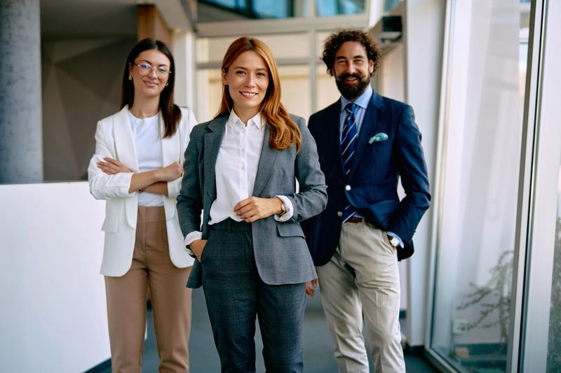 Business team of three people smiling confidently at camera, standing together in a modern brightly lit office corridor, portraying corporate success and professionalism