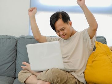 Young man celebrating joyfully while using a laptop on a couch.