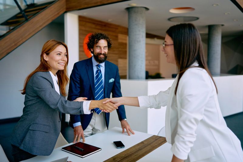 Two smiling businesswomen shaking hands during an office meeting, with a businessman smiling in the background, symbolizing partnership, agreement, and a new deal