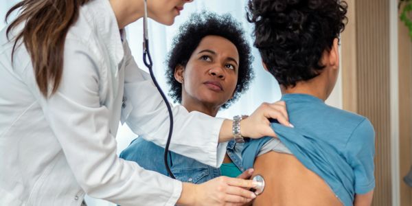 Doctor using a stethoscope to check a boy's lungs while his mother watches.