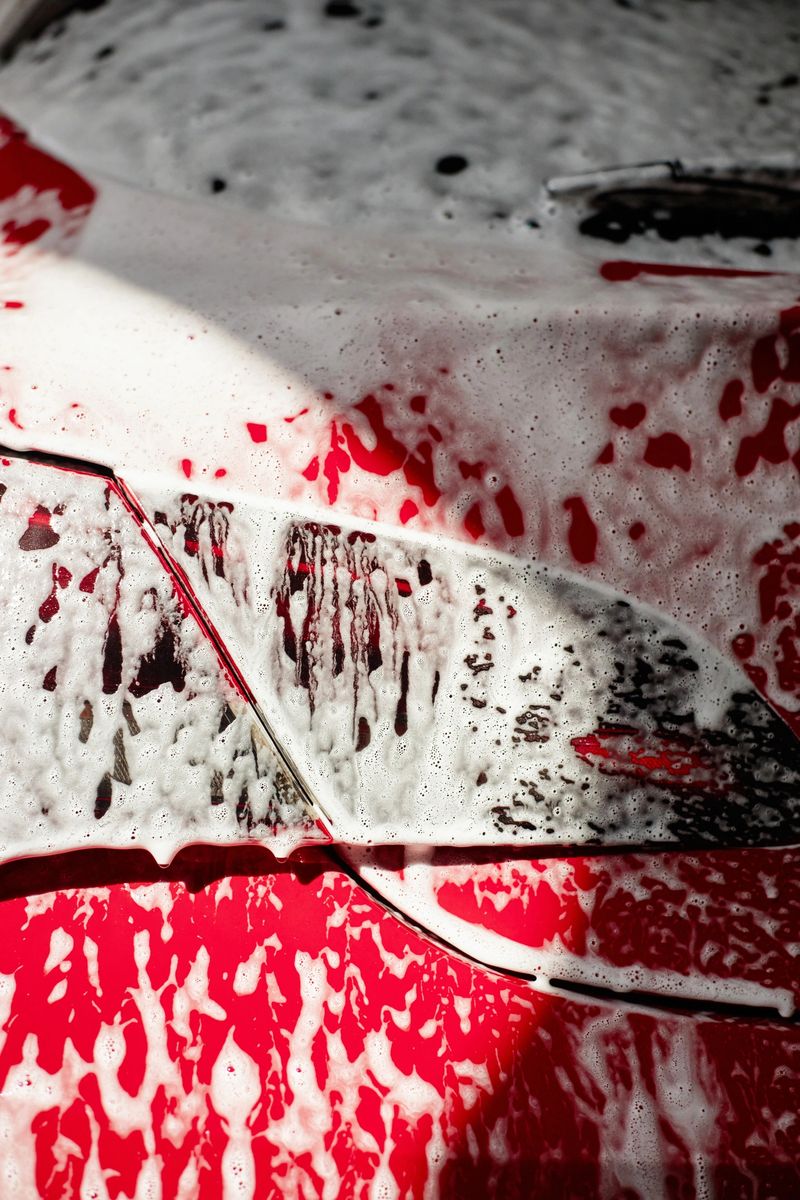 Closeup showing red car hood covered in thick soap suds during washing process, foam and water streaming down glossy surface, sunlight casting shadows across vehicle
