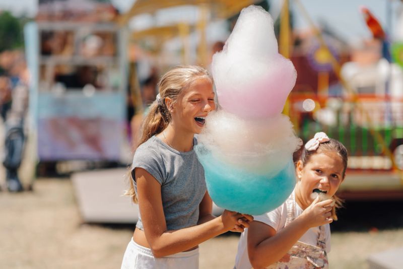 Two sisters are having fun eating a huge, colorful cotton candy at an amusement park, creating a sweet and joyful summer memory