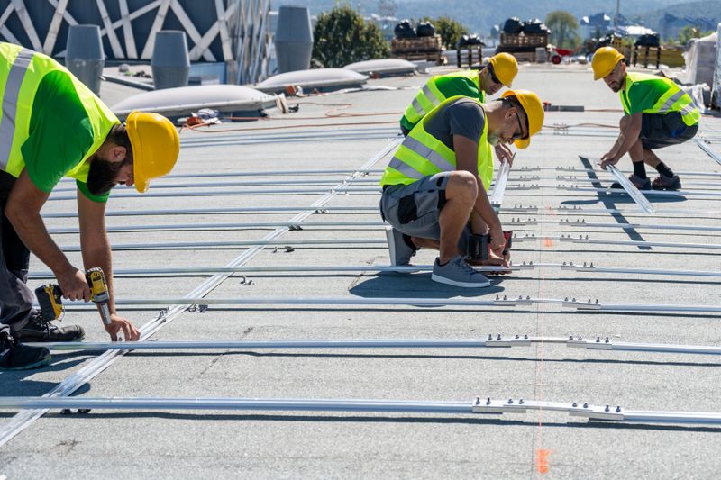 Group of technicians working on solar panel setup featuring rooftop installation. Wearing safety vests and helmets, they secure mount rails using drills, demonstrating teamwork and focus. Outdoor setting with clear sky and urban background.