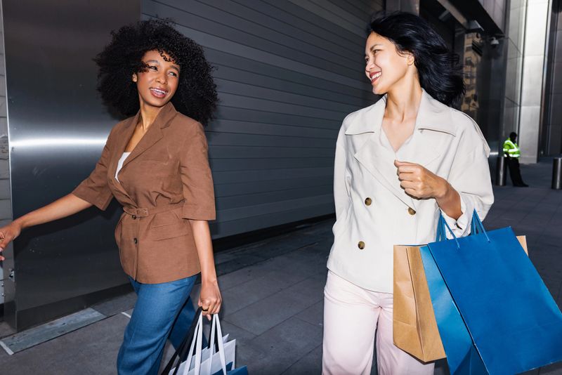 Two stylish women walk together in an urban setting, smiling and chatting while carrying multiple colorful shopping bags. The scene captures friendship, fashion, and the joy of a city shopping day.