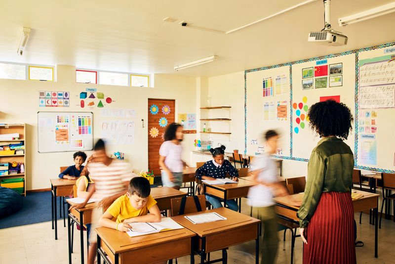 Female teacher guiding a diverse group of schoolchildren during class in a classroom decorated with colourful charts and educational posters.