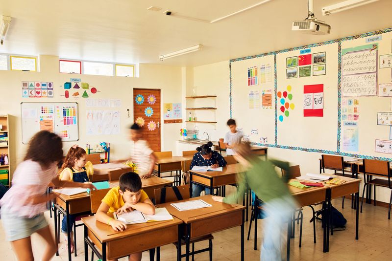 Energetic students engaging between desks in a bright classroom filled with sunlight and colourful wall charts, showing the vibrancy, movement, and social energy of real school life.