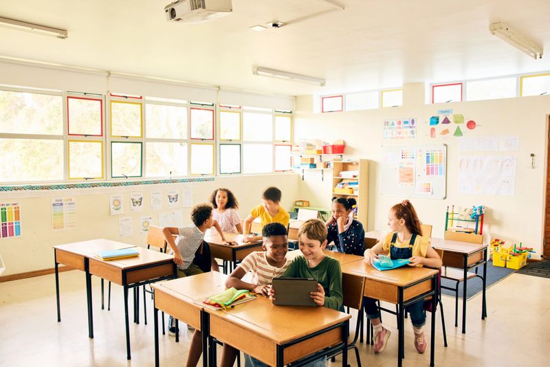 Diverse group of schoolchildren collaborating in a colourful classroom, sharing a digital tablet and writing in notebooks, reflecting teamwork, learning, and modern education.