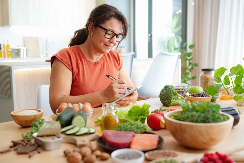 Woman sitting at the table full of healthy ingredients for MIND diet. Food to reduce dementia and brain failure. Planning a healthy-brain diet, balanced food or healthy living. Mediterranean diet