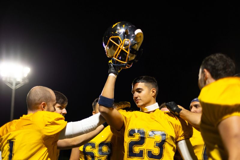 American football player holding up helmet, celebrating a team victory. Teammates congratulating him on the field under stadium lights