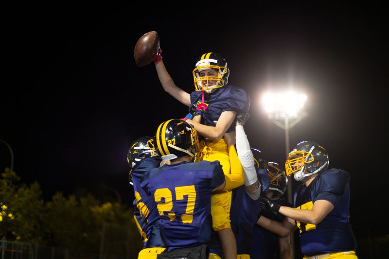 Youth american football team celebrating victory on the field at night, lifting a smiling player holding a football