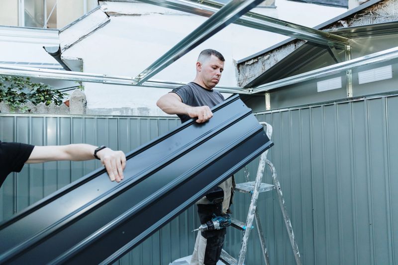One Caucasian young worker standing on a stepladder and taking steel roofing sheets from his assistant to install on a metal shed in the backyard on a summer day, side view, medium shot. Concept assembly metal shed, time teamwork, using technology.