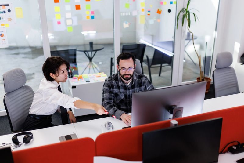 Two business colleagues are working together at a desk in a bright, modern office. A woman is pointing at the computer screen while the man focuses on the task at hand.