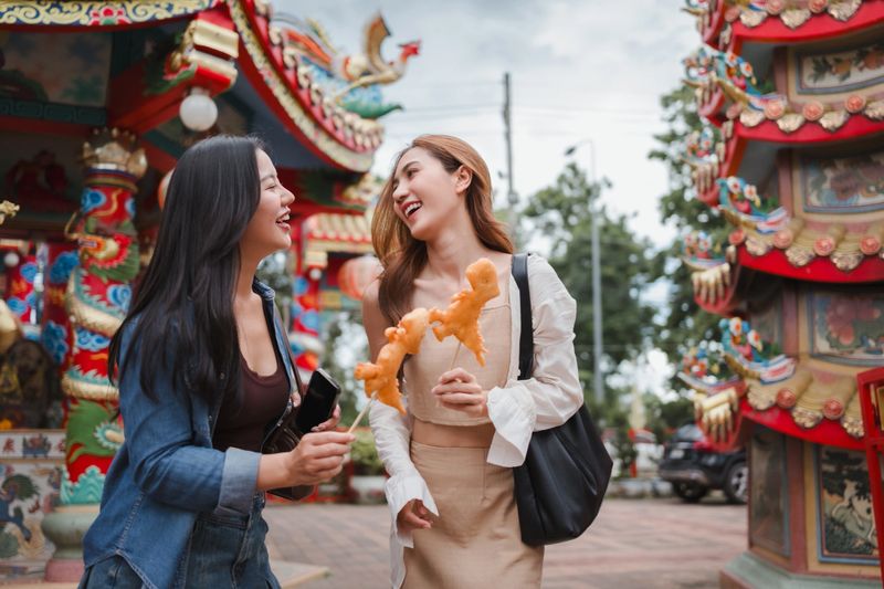 Two young adult women laughing, holding street food outside a Chinese temple
