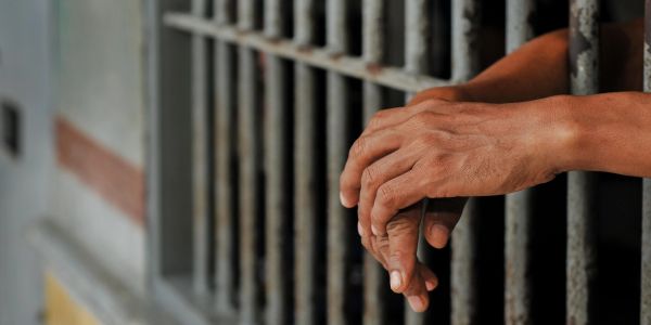 Hands clasped behind prison bars, conveying confinement and isolation.