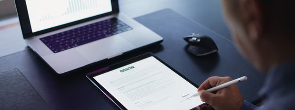 Person signing a digital contract on a tablet with a laptop nearby.