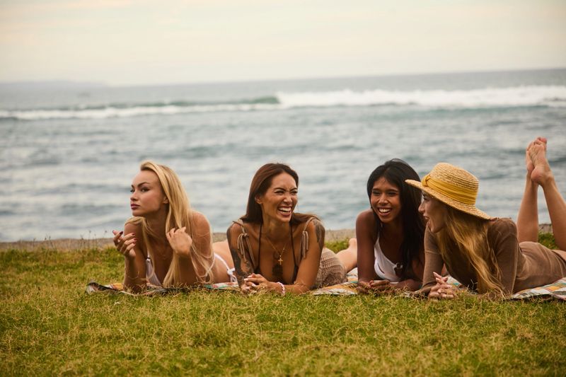 A group of four women relaxes on a grassy spot by the ocean, smiling and talking together on a sunny day. They wear casual beachwear, creating a warm, carefree moment.
