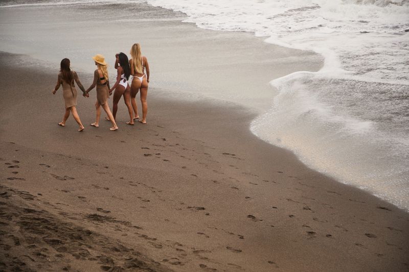 Five women walk hand in hand along a sandy beach, wearing swimsuits and summer dresses, as gentle waves wash onto shore. A relaxed, carefree, vacation mood with friendship and togetherness.