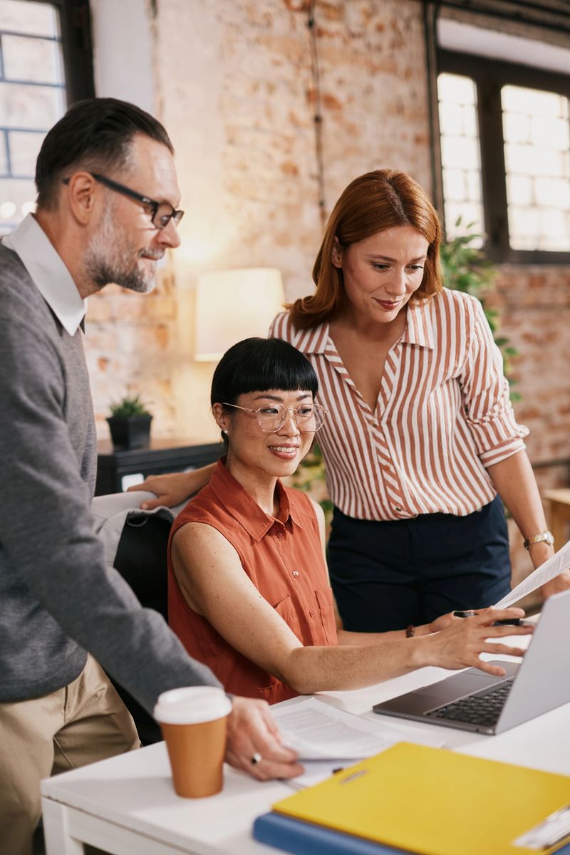 Three adult, mature colleagues collaborate around a white desk in a modern loft, one seated at a laptop while two stand, reviewing documents. A warm, supportive, focused atmosphere fosters teamwork, creativity, and problem solving.