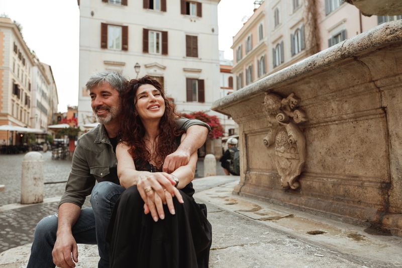 A mature couple relaxes beside a historic fountain in Rome’s charming Piazza Madonna dei Monti. The man embraces his smiling partner as they soak in the bohemian atmosphere of the Monti district, surrounded by baroque architecture and lively cafés—capturing the authentic essence of Italian tourism.