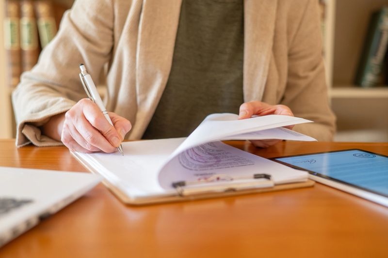 Woman's hands signing a contract on a clipboard while reviewing additional financial documents, indicating a successful business deal, loan approval, or real estate transaction