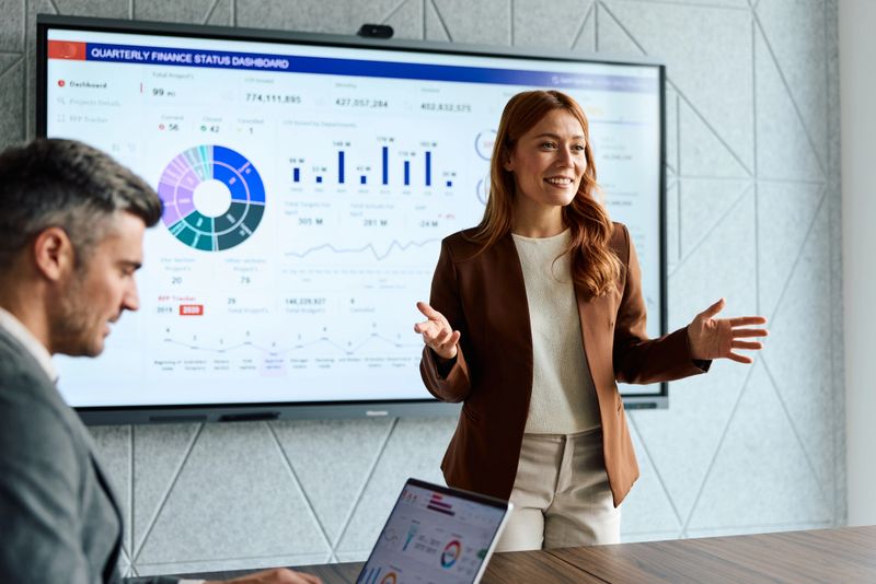 Confident businesswoman talking and gesturing during a corporate presentation in a contemporary office, showcasing financial analysis on a large digital screen while a colleague works on a laptop