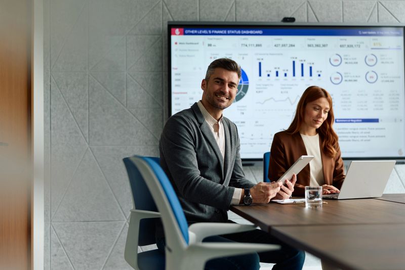Two business professionals reviewing finance dashboard on large screen and digital devices, collaborating on data analysis during an important corporate meeting
